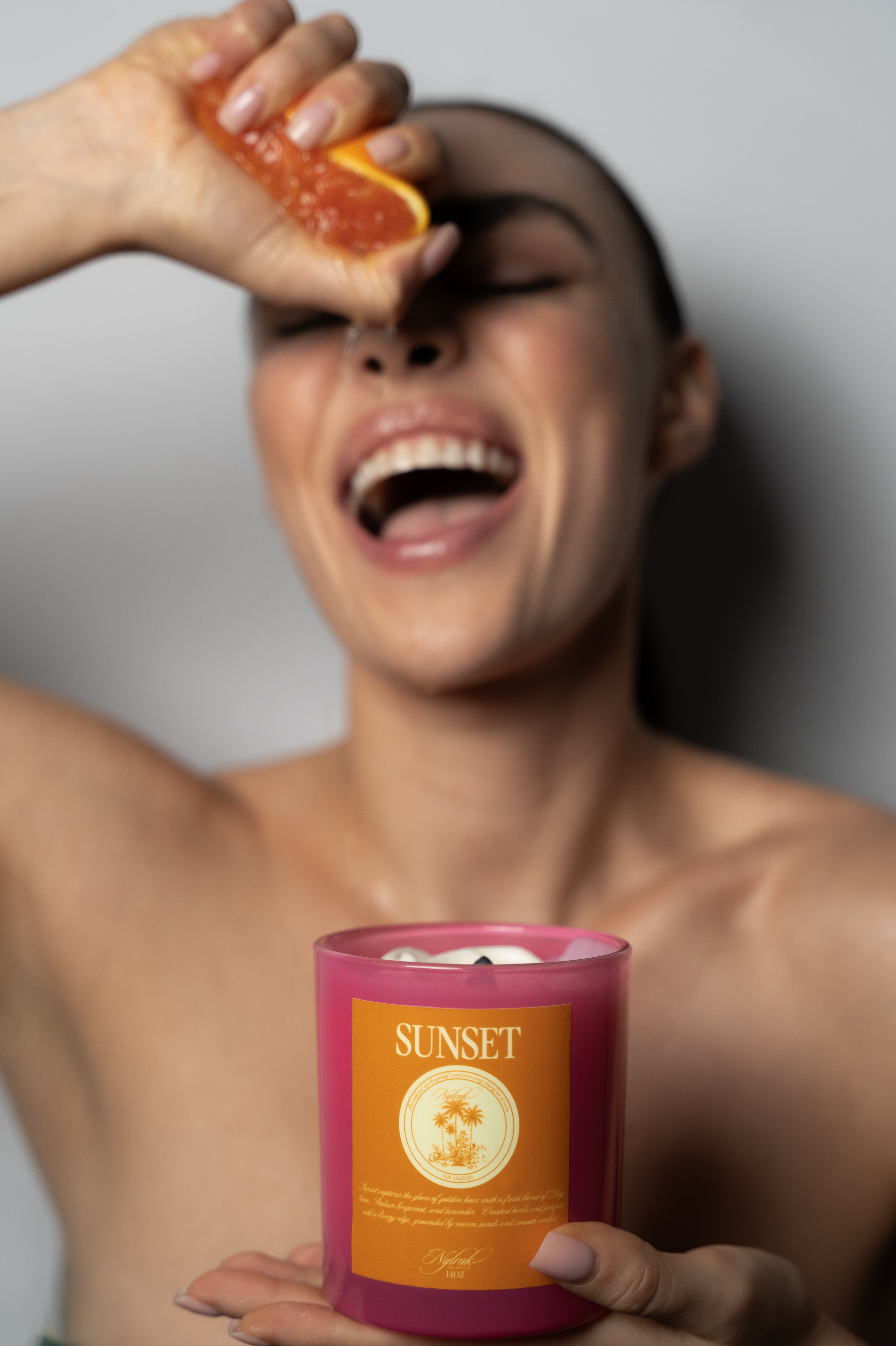 Woman holding a Nylrak 'Sunset' candle  with a slice of fruit above her eye.